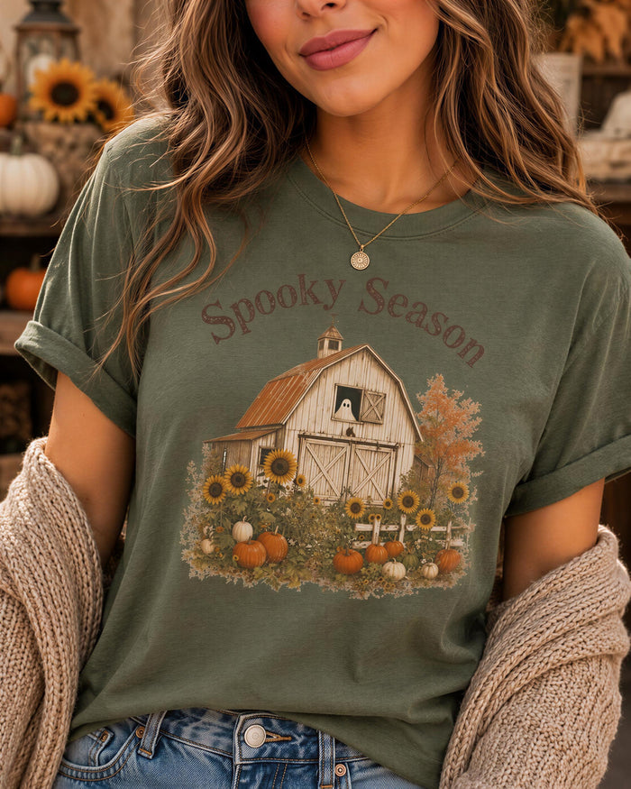 A woman wearing a green t-shirt with a graphic of a barn and autumn foliage, along with the text "Spooky Season".