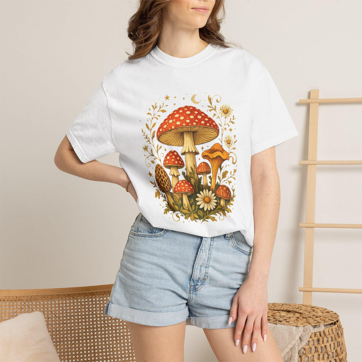 A woman wears a white short-sleeved t-shirt with a graphic of red and orange mushrooms, white flowers, and gold leaves, paired with light blue denim shorts.