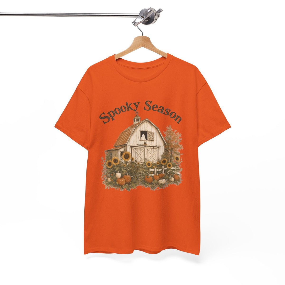 An orange t-shirt hanging on a wooden hanger, displaying a graphic of a white barn, sunflowers, and pumpkins, with "Spooky Season" text above.
